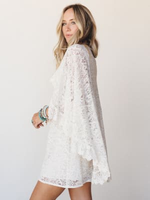 An ivory lace cape dress with dramatic bell sleeves and scalloped hem worn by a blonde model posed three-quarter away from the camera — romantic and relaxed with a stack of turquoise and gold bracelets adding just the right amount of personality.