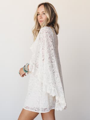 An ivory lace cape dress with dramatic bell sleeves and scalloped hem worn by a blonde model posed three-quarter away from the camera — romantic and relaxed with a stack of turquoise and gold bracelets adding just the right amount of personality.