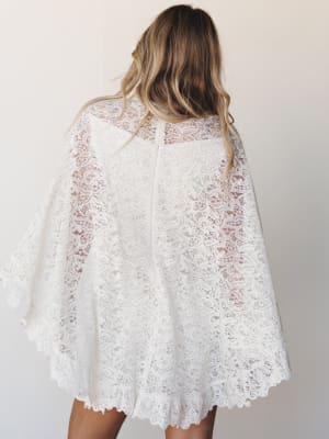 A back view of an all-over ivory lace cape dress with dramatic wide sleeves and a scalloped hem — romantic and dreamy, like something you'd wear to say 'I do' or dance barefoot at a garden party.