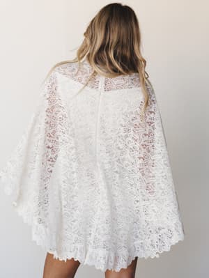 A back view of an all-over ivory lace cape dress with dramatic wide sleeves and a scalloped hem — romantic and dreamy, like something you'd wear to say 'I do' or dance barefoot at a garden party.