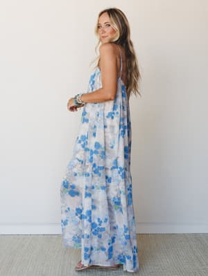 A flowy floral maxi dress in soft blue and white shot against a clean studio wall — the kind of dress that makes you want to book a trip somewhere warm immediately.