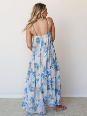 Shot from the back, a flowy tiered maxi dress in a soft blue floral print drapes all the way to the floor — the kind of dress that looks equally good at a summer wedding or wandering a farmers market.