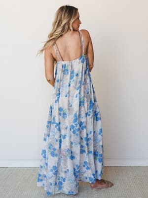 Shot from the back, a flowy tiered maxi dress in a soft blue floral print drapes all the way to the floor — the kind of dress that looks equally good at a summer wedding or wandering a farmers market.
