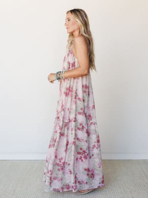 A flowy floral maxi dress in soft mauve and rose tones photographed in profile — tiered and airy, styled with stacked turquoise bracelets for that effortless boho finish.