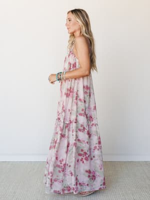 A flowy floral maxi dress in soft mauve and rose tones photographed in profile — tiered and airy, styled with stacked turquoise bracelets for that effortless boho finish.