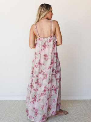 A flowy floral maxi dress photographed from the back, showing off its open back, adjustable spaghetti straps, and tiered skirt — soft rose print on a blush ground that feels like something you'd wear to a garden party and never want to take off.