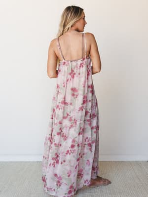 A flowy floral maxi dress photographed from the back, showing off its open back, adjustable spaghetti straps, and tiered skirt — soft rose print on a blush ground that feels like something you'd wear to a garden party and never want to take off.