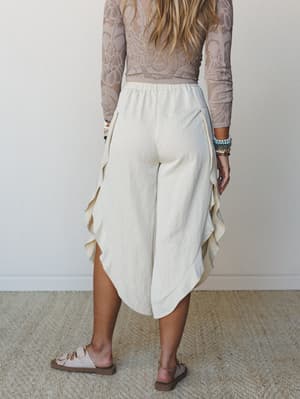 Shot from behind, she's wearing flowy linen-blend wide-leg pants with ruffled side hems paired with a lace long-sleeve top — the kind of effortless outfit that looks just as good on a beach boardwalk as it does at a weekend brunch.