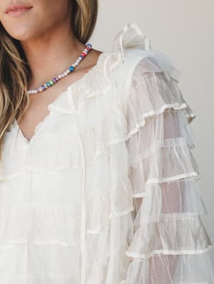 A close-up detail shot of an ivory tulle blouse with tiered ruffle sleeves and a ruffled neckline, styled with a colorful beaded pearl necklace — romantic and a little dreamy.