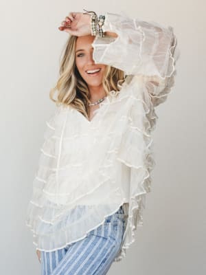 A smiling blonde model in a dreamy ivory ruffle blouse with sheer layered tiers paired with striped wide-leg jeans, shot against a clean studio wall with a relaxed, joyful energy.