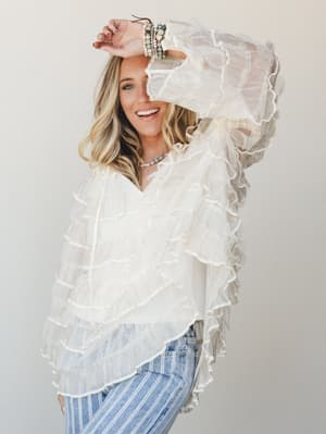 A smiling blonde model in a dreamy ivory ruffle blouse with sheer layered tiers paired with striped wide-leg jeans, shot against a clean studio wall with a relaxed, joyful energy.