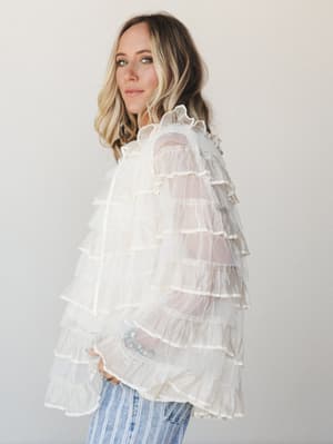 A dreamy tiered ruffle blouse in sheer ivory mesh shot against a soft neutral studio wall — the kind of top that makes jeans feel like a whole moment.