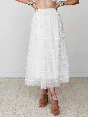 A dreamy ivory tulle maxi skirt with layered horizontal ruffles, styled with a cropped top, stacked beaded bracelets, and tan suede platform clogs — the kind of outfit that makes you feel like you wandered out of a garden party.