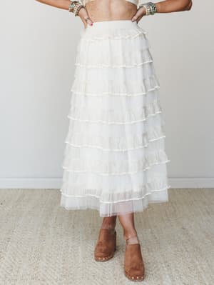 A dreamy ivory tulle maxi skirt with layered horizontal ruffles, styled with a cropped top, stacked beaded bracelets, and tan suede platform clogs — the kind of outfit that makes you feel like you wandered out of a garden party.
