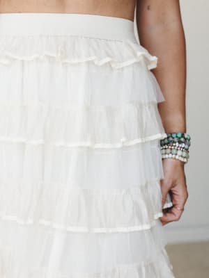 A close-up of an ivory tiered tulle ruffle skirt with a wide elastic waistband, styled with a stack of beaded stone bracelets — soft, romantic, and full of texture.