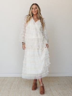 A flowy ivory tiered ruffle maxi dress with sheer long sleeves worn with tan suede clogs and stacked turquoise bracelets — romantic and a little dreamy, like she's already running late to the garden party.