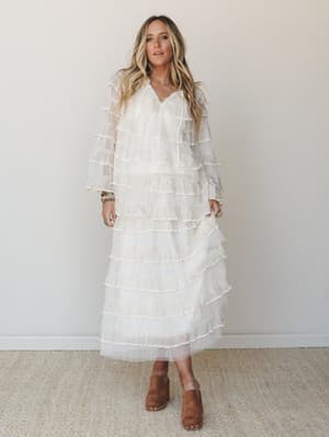 A flowy ivory tiered ruffle maxi dress with sheer long sleeves worn with tan suede clogs and stacked turquoise bracelets — romantic and a little dreamy, like she's already running late to the garden party.