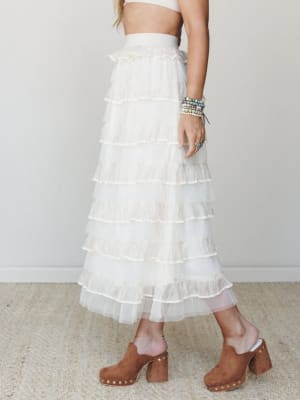A dreamy ivory tiered ruffle maxi skirt styled with a cropped top and brown suede studded clogs — the kind of outfit that makes a bridal shower feel effortless and a festival feel fancy.