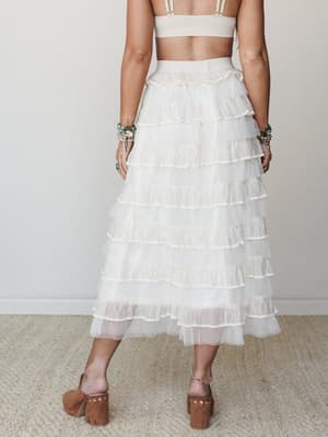 Shot from the back, a model wears a dreamy ivory tiered ruffle midi skirt paired with a cream crop top, stacked turquoise bracelets, and suede block-heel clogs — the whole look has that effortless something-borrowed-something-boho energy.