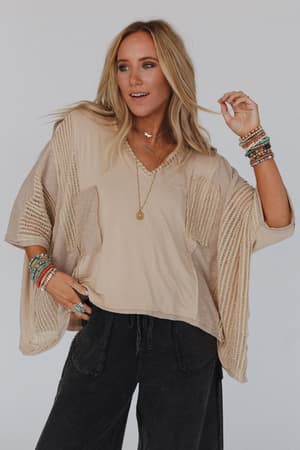 A woman with long blonde hair wears the City Drifter Oversized Contrast Top in taupe, paired with dark pants and vibrant accessories, creating a relaxed boho vibe against a plain light background.