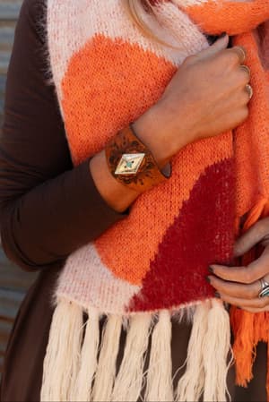 A person wears a cozy, orange and red patterned scarf, a brown long-sleeve top, and the Wynona Diamond Leather Cuff Bracelet in camel with turquoise accents—holding the scarf: a perfect boho accessory.