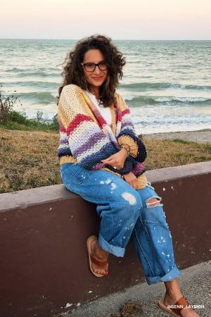 Woman wearing a colorful striped sweater and blue jeans sitting by the ocean.