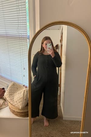 A woman stands barefoot in front of a tall, curved mirror taking a selfie. She wears the Simply Comfort Jumpsuit - Black. Beside her are a quilt and wicker basket, and blinds cover the window behind her.
