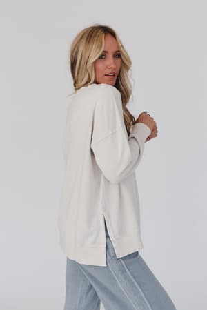 A woman with long blonde hair wears the Catch My Smile Pullover Top in khaki and light blue pants, standing sideways and looking over her shoulder against a plain light background.