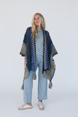 A woman with long blonde hair wears the Desert Dreams Crochet Ruana – Cobalt from her boho closet over a light blue outfit and white sneakers, standing against a plain white background.
