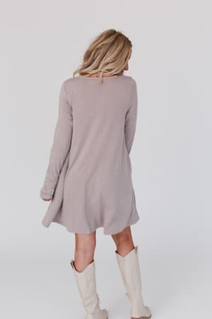 A woman with blonde hair wears The Nest Jake Long Sleeve Crewneck Dress in Mocha Latte and cream cowboy boots, standing facing away against a plain white background.