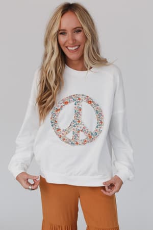 A woman with long blonde hair smiles against a plain light background, wearing the Jenny Floral Peace Sign Pullover Top in off white and orange pants.