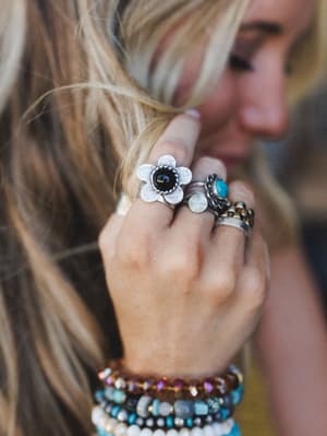 A close-up of stacked boho rings and layered beaded bracelets on a model's raised hand, with a silver flower ring set with a black stone taking center stage — the kind of jewelry stack that looks collected over time, not bought all at once.