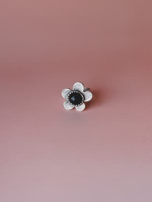 A silver flower-shaped ring with a brushed metal finish and a dark black stone center, shot against a warm dusty rose background — the kind of piece that quietly makes every outfit feel a little more intentional.