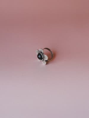 A silver flower-shaped ring with a dark cabochon stone center sits against a dusty rose gradient background — the kind of piece that looks like it's been in someone's jewelry box since a really good summer.