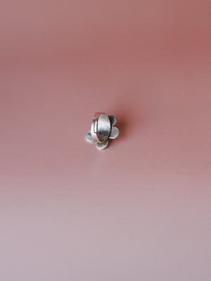 A small silver flower-shaped ring photographed from above against a muted dusty rose background — simple, handcrafted-feeling, and quietly sweet.