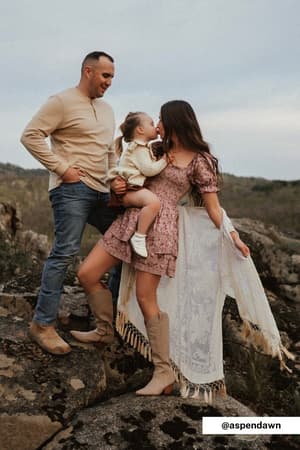 A man, woman, and young child stand on outdoor rocks. The woman wears the Crushin' On You Velvet Kimono - Nude as she holds the child, who kisses her cheek. The man in jeans and a beige shirt smiles at them, with hills and greenery in the background.