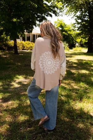 A woman with long, wavy hair stands on grass outdoors, facing away. She wears the Way Home Patch Detail Shacket - Beige featuring a large white lace mandala on the back, loose blue jeans, and beige flats in filtered sunlight.