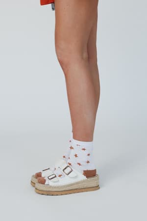 A person wears Super Star Socks - White, featuring an orange star pattern, styled with white sandals with thick soles and buckles. The standout socks create a bold look against a plain light background.