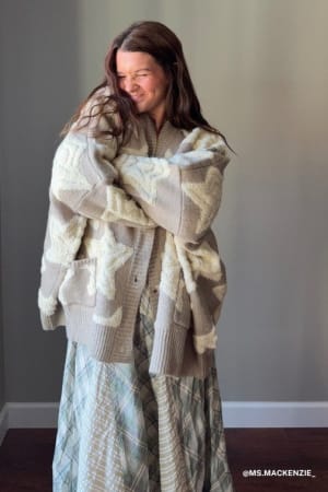 A woman with long brown hair smiles with her eyes closed, hugging herself while wearing the Wrapped In Stars Oversized Cardigan in taupe over a patterned skirt, standing against a plain gray wall.