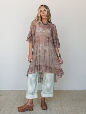 A sheer taupe lace tunic with ruffle trim and flutter sleeves is layered over mint gingham wide-leg pants and brown clogs — the kind of effortlessly mixed outfit that looks accidental but absolutely isn't.