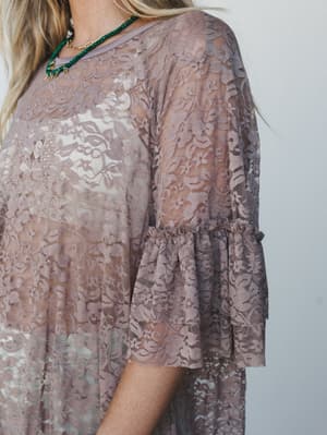 A close-up detail shot of a flowy lace tunic in soft taupe, showing the delicate floral lace overlay, ruffle-trimmed bell sleeves, and a pop of emerald green beaded necklace at the neckline.
