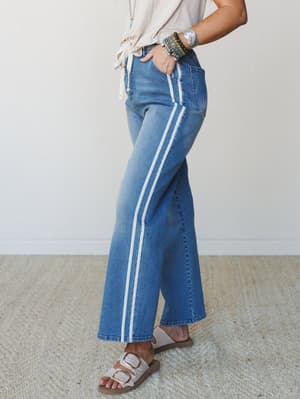 A side-profile shot of wide-leg medium wash jeans with a double white side stripe, styled with a knotted cream tee, stacked boho bracelets, and buckle sandals — easy, retro-leaning, and very much a Saturday kind of outfit.