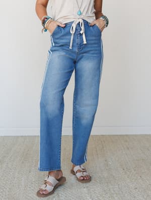 Medium wash straight-leg jeans with a cream drawstring waist and side stripe detail, styled with a knotted tee, stacked bracelets, and buckle sandals for an easy, expressive everyday look.