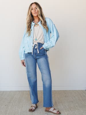 A relaxed full-body shot of a model in medium wash straight-leg jeans with white side stripes, layered under a powder blue cropped jacket and a neutral tank — the kind of effortless outfit that looks like it took ten minutes but really took instinct.