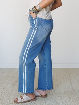A side-profile shot of wide-leg medium wash jeans with double white side stripes, styled with a cream tie-front top, beaded bracelets, and neutral buckle sandals — relaxed and a little retro in the best way.