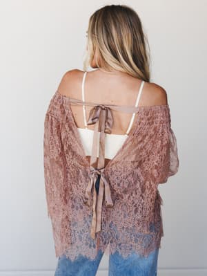 Shot from behind, a model wears a dusty mauve lace tunic with an off-shoulder neckline and dramatic bell sleeves, styled open-back with satin ribbon tie closures over a white bralette and jeans — romantic and a little undone in the best way.