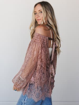 A dusty rose lace off-the-shoulder tunic with billowy sleeves and a satin ribbon tie at the back, styled over a white cami and blue jeans — romantic and a little undone in the best way.