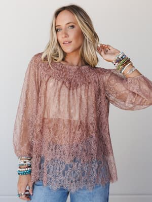 A dusty mauve lace tunic with smocked neckline and scalloped hem, styled with stacked beaded bracelets and jeans — romantic and a little undone in the best way.