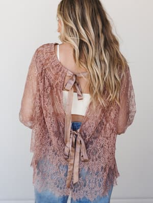 Shot from behind, a lace kimono in dusty rose drapes open over a white crop top and denim, tied at the back with two satin ribbon bows — the kind of piece that makes a casual outfit feel like a whole moment.