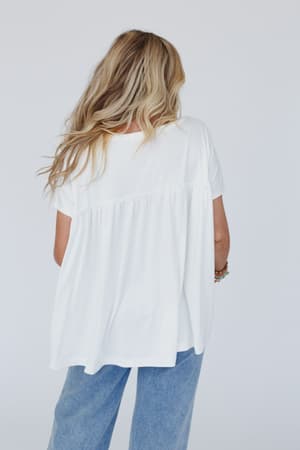 A person with long, wavy blonde hair models the Sunday Morning Babydoll Top in white and light blue jeans, standing with their back to the camera against a plain light background.